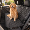 A golden retriever sitting on a black quilted dog car seat cover on a car back seat, with a brick house and hedge through the window